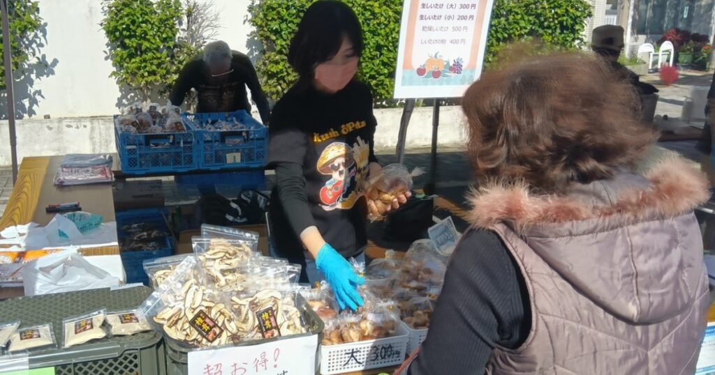とらいあんぐるカフェのお店でとらいあんぐる農園の乾燥椎茸を手に取る来場者と、説明を行うスタッフ。安佐公民館まつりでの販売の様子。