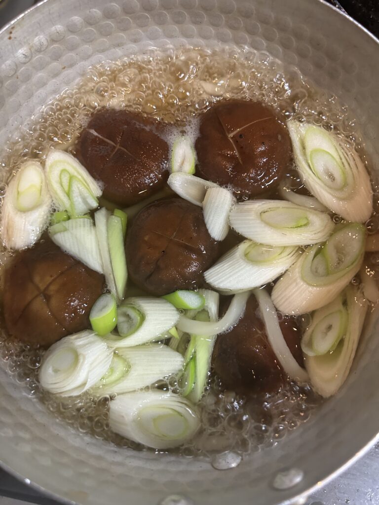 輪切りにした白ネギと肉厚椎茸を煮込んでいる様子。出汁がぐつぐつと煮立ち、旨みが広がるシーン。
