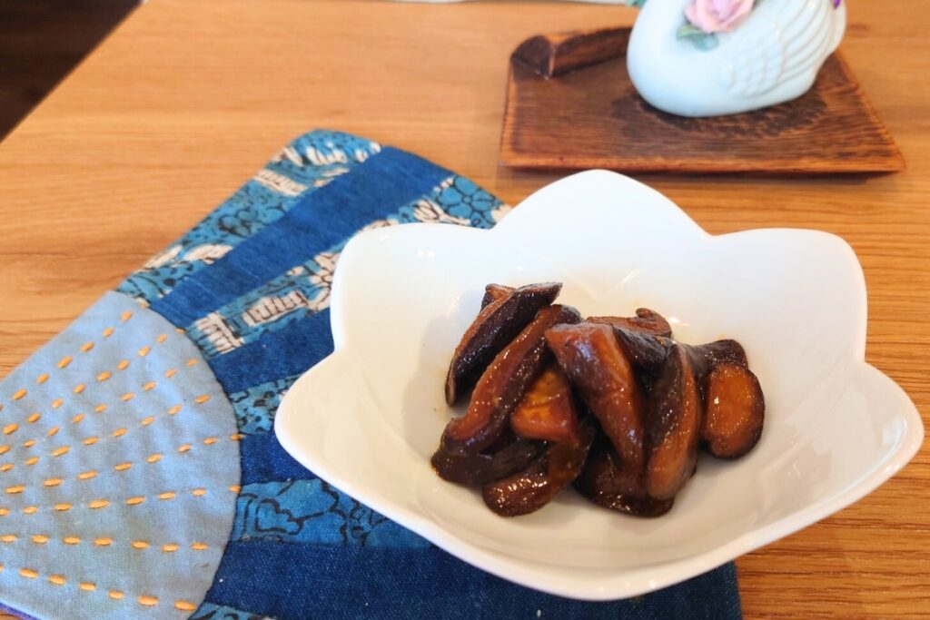 白い花型の器に盛りつけられた椎茸の辛子和え。木のテーブルの上に、藍色の和柄の布とともに置かれている。とらいあんぐる農園の手作りレシピ。