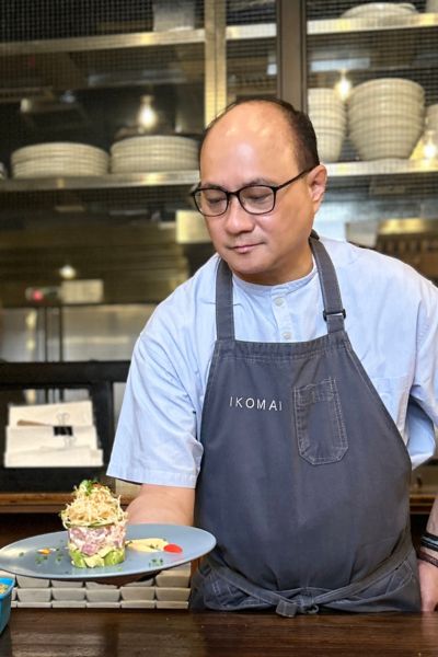 IKOMAIオーナーのジェームス・アントリンさんが、レストランカウンターで美しく盛り付けられた料理を手にしている様子。胸元には「IKOMAI」のロゴ入りエプロン。